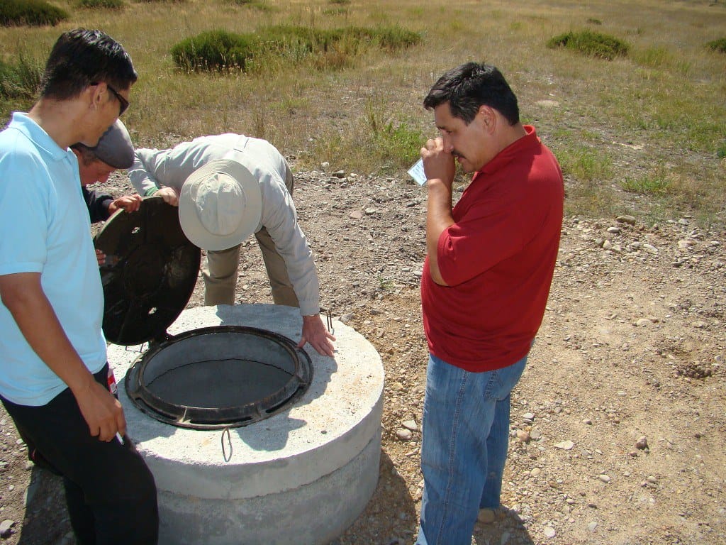 Four individuals gathered around a septic tank opening in a rural area.