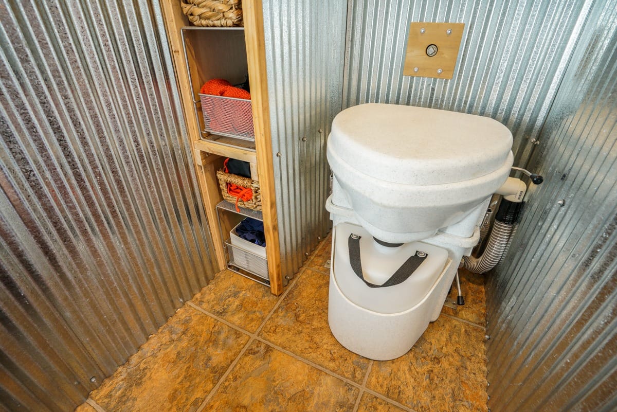 Waterless toilet installed in a small bathroom with metal walls and storage shelves.