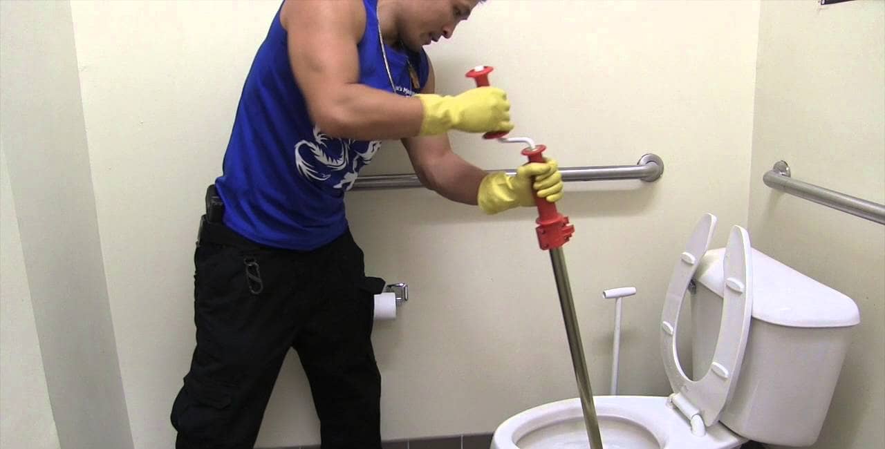 Individual wearing gloves using a toilet plunger to unclog a toilet in a restroom.