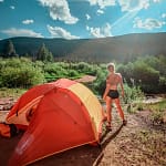 A person standing near an orange camping tent in a natural outdoor setting under a bright sky.