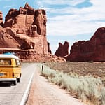Yellow Volkswagen van driving on a road surrounded by red rock formations in a desert landscape.