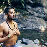 topless man wearing black beaded necklace and blue denim shorts standing on rocky shore during daytime