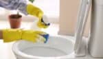 Person wearing yellow gloves cleaning a toilet bowl with a sponge and cleaning solution.