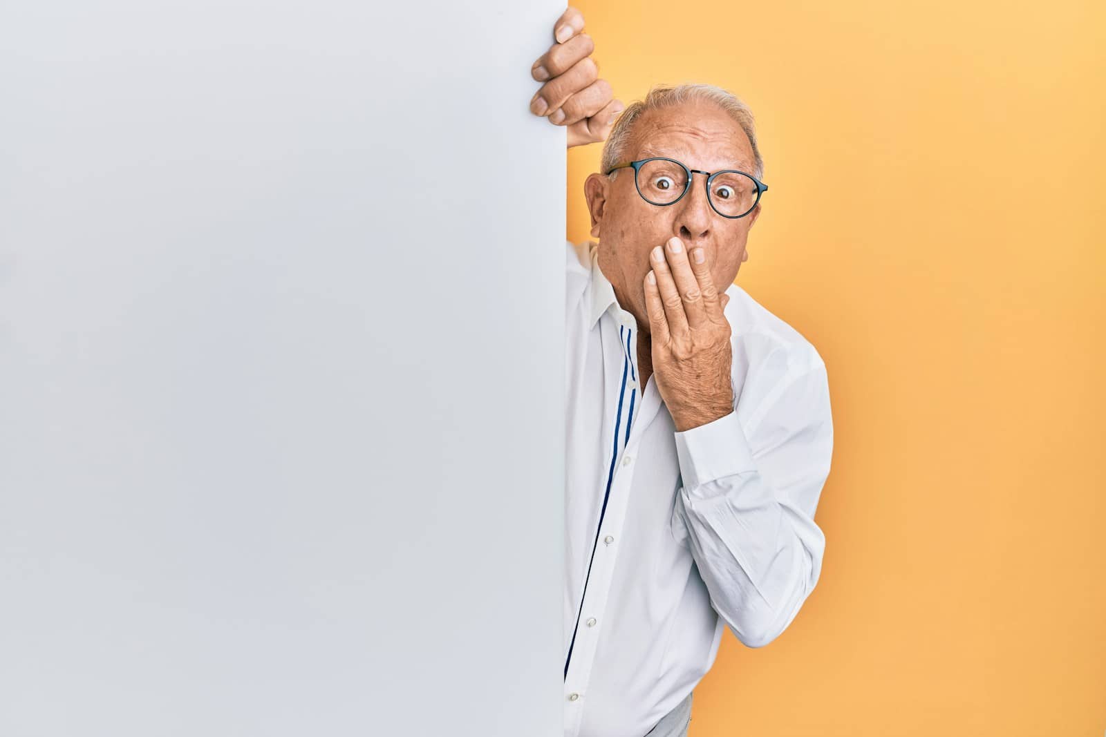 Elderly man with glasses and a white shirt peeking around a blank panel with a surprised expression.