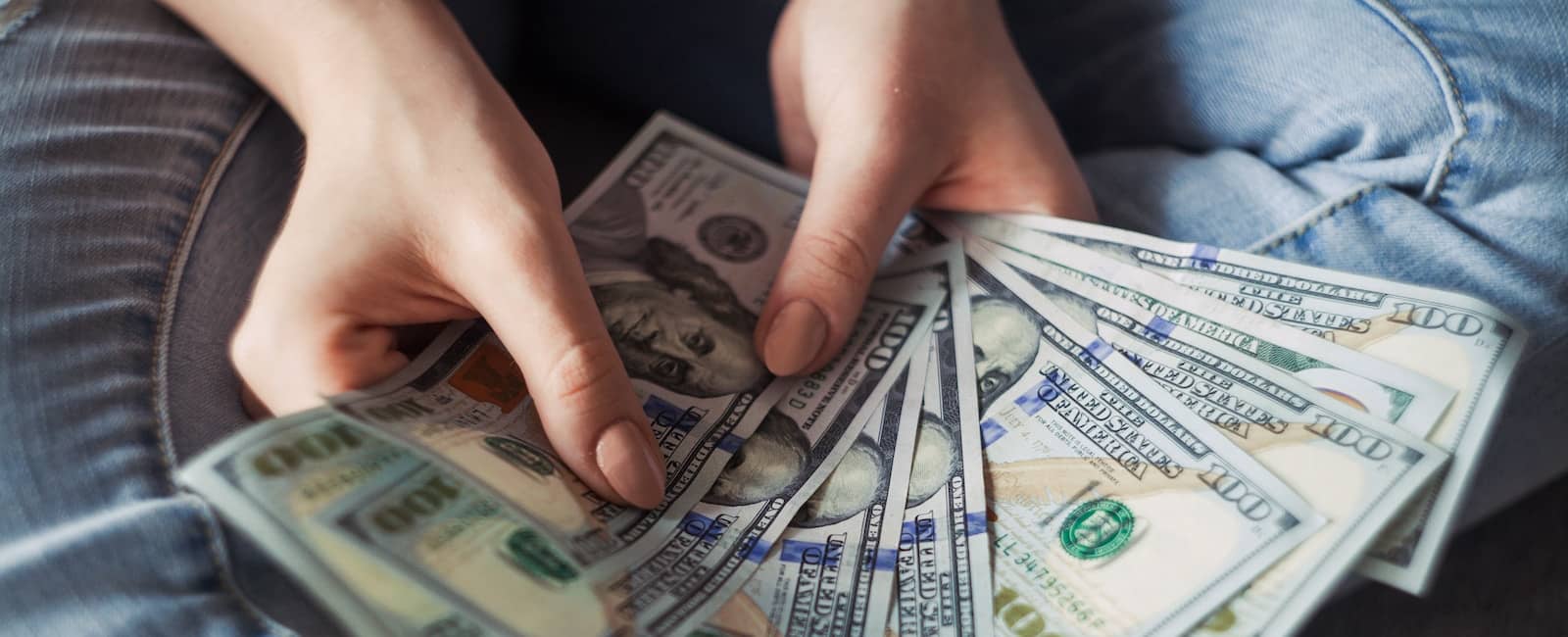 Individual holding a fan of hundred dollar bills in both hands, showing the front side of the currency.