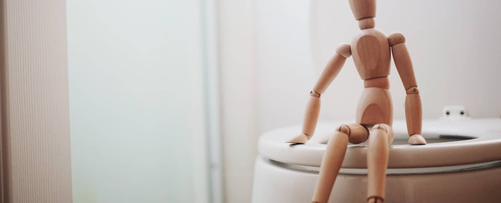 Wooden articulated figure sitting on the rim of a toilet in a bathroom setting.
