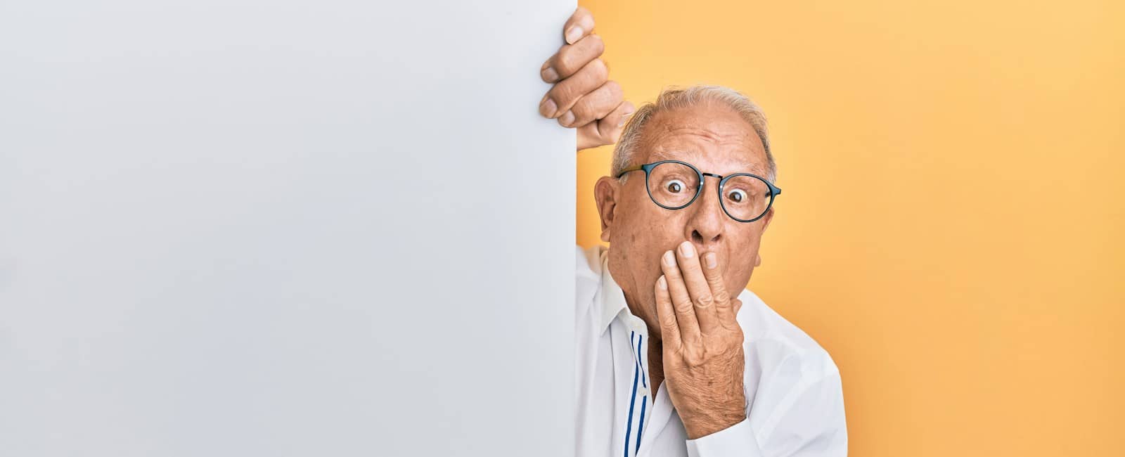 Elderly man with glasses and a white shirt peeking around a blank panel with a surprised expression.