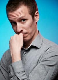 A man with short hair and a gray shirt, resting his chin on his hand with a thoughtful expression.