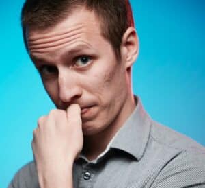 A man with short hair wearing a gray shirt, looking thoughtfully with his hand near his mouth.