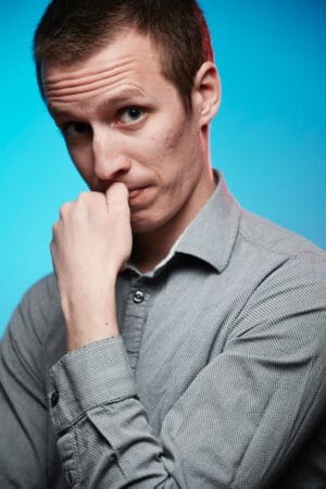 A man with short hair wearing a gray shirt, looking thoughtfully with his hand near his mouth.