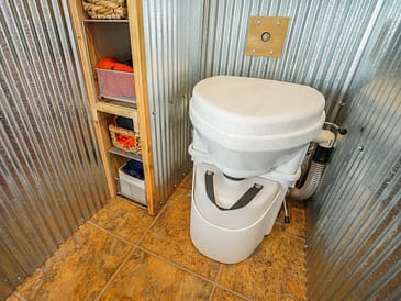 Waterless toilet installed in a small bathroom with metal walls and storage shelves.