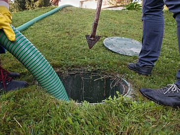 A worker using a green striped hose to service a septic system access point in a grassy area.