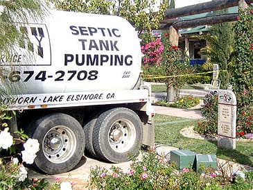 A woman standing near a septic tank pumping truck in a landscaped area with flowers.