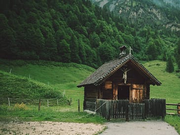 A small wooden chapel with a cross on top, surrounded by trees and grass.