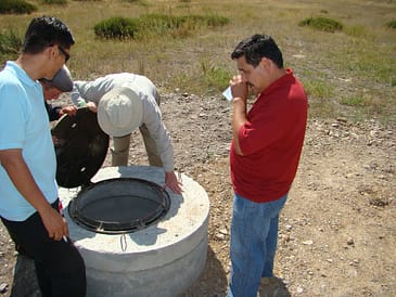 Four individuals gathered around a septic tank opening in a rural area.