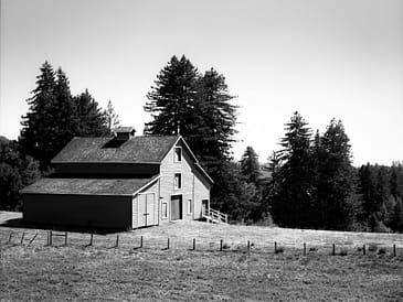 Black and white image of a barn with a sloped roof and trees in the background.
