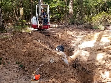 Mini excavator operating at an excavation site in a wooded area with exposed soil.