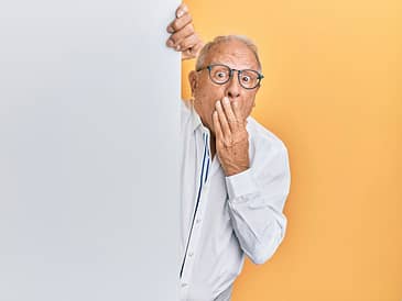 Elderly man with glasses and a white shirt peeking around a blank panel with a surprised expression.