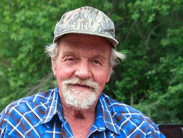 Older man wearing a blue plaid shirt and a camouflage cap, smiling at the camera.