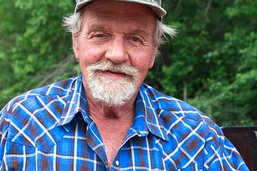 Older man wearing a blue plaid shirt and a camouflage cap, smiling at the camera.