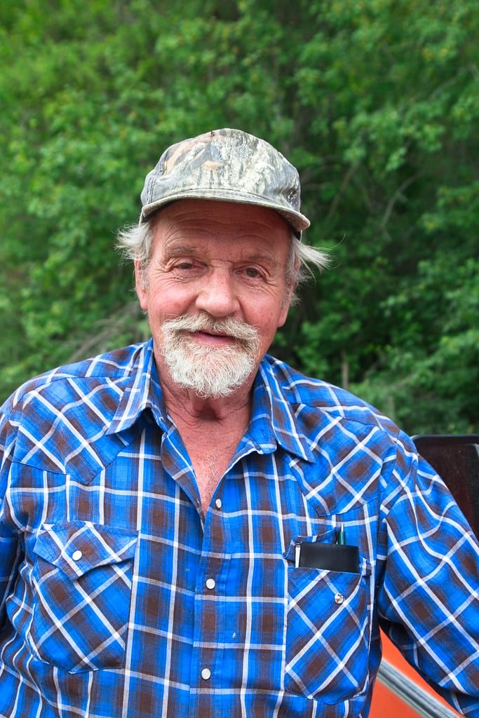 Older man wearing a blue plaid shirt and a camouflage cap, smiling at the camera.