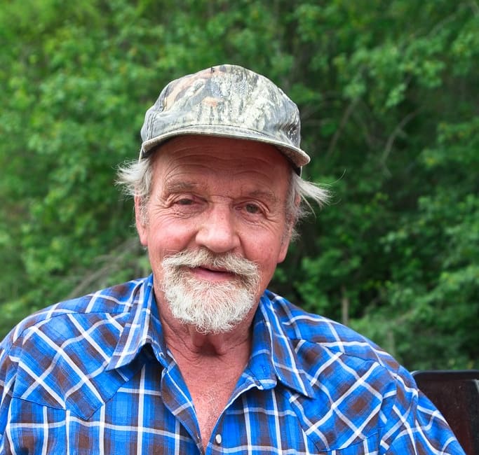 Older man wearing a blue plaid shirt and a camouflage cap, smiling at the camera.