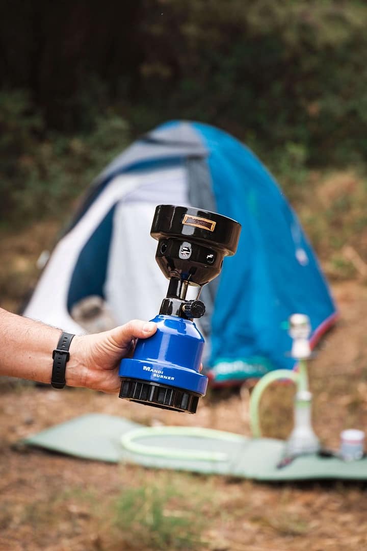 Hand holding a blue portable camping stove with a black top in a natural outdoor setting.