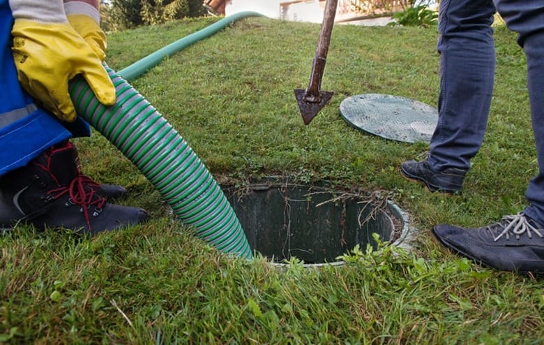 A worker using a green striped hose to service a septic system access point in a grassy area.